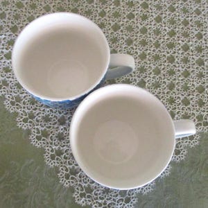 May include: Two white ceramic mugs with light blue handles and a blue patterned design around the rim. The mugs are empty and sit on a green surface with a white lace pattern.