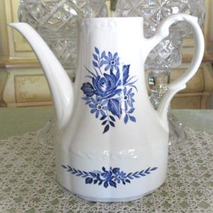 May include: White ceramic coffee pot with a long spout and a curved handle. The pot is decorated with blue floral designs, including a large rose and smaller flowers. A blue floral garland adorns the base of the pot.