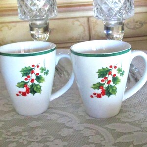 May include: Two white ceramic mugs with a green rim and handle. Each mug features a holly and berry design in red and green. The mugs are on a patterned surface.