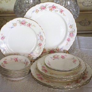 May include: A collection of white porcelain dinnerware with a floral rose pattern. The set includes plates of various sizes and small bowls. Each piece has a gold trim and scalloped edges. The floral design features pink, yellow, and blue roses.