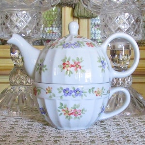 May include: White porcelain tea-for-one set with a teapot and teacup. The set features a floral pattern in red, purple, and yellow. The teapot has a lid and a curved spout. The teacup has a handle.