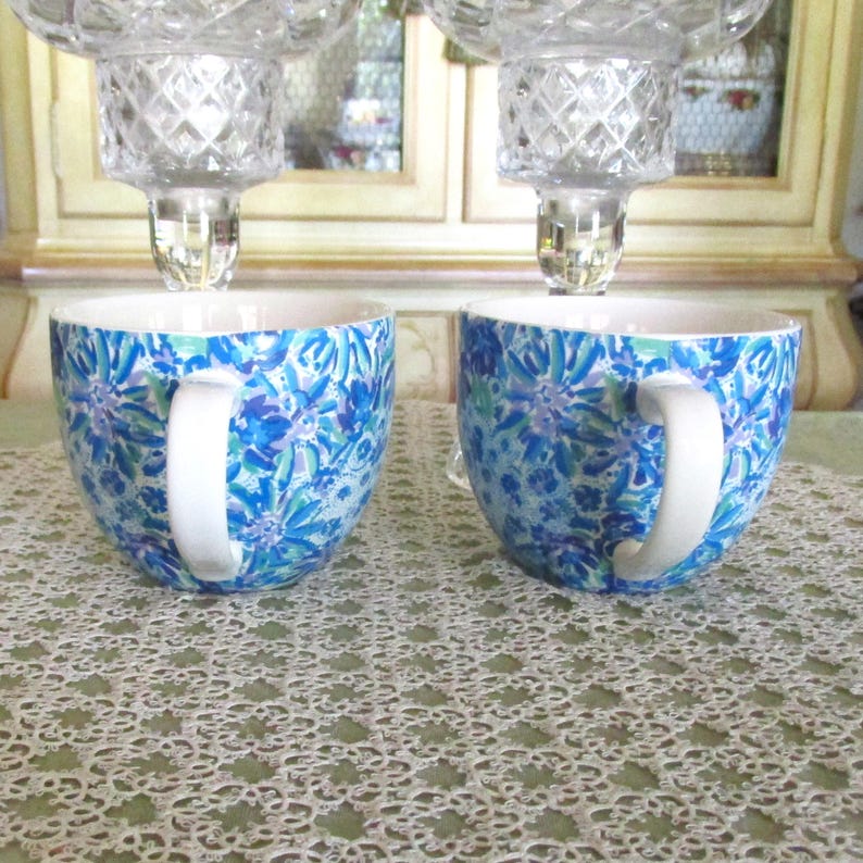 May include: Two ceramic mugs with white handles and a blue floral pattern. The mugs are placed on a white lace tablecloth. Above the mugs are two clear crystal candle holders.