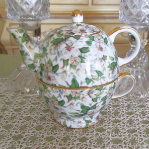 May include: A white porcelain teapot with a floral design of white flowers and green leaves. The teapot has a gold trim around the lid and the base. The teapot is a double-walled design with a built-in cup.