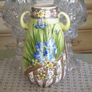 May include: A green and white ceramic vase with two handles. The vase is decorated with blue and gold irises and other flowers. The vase has a gold rim and a gold band around the middle.