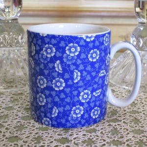 May include: A ceramic mug with a white handle and interior. The exterior features a blue floral pattern with white flowers and green leaves. The mug is designed for hot beverages.