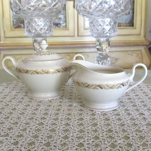 May include: Cream-colored ceramic sugar bowl with lid and creamer set. Both pieces have a gold leaf pattern around the rim. Two crystal candle holders are in the background, and the set is on a lace tablecloth.