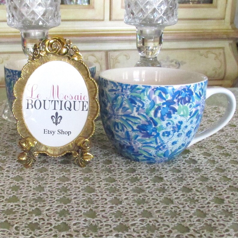 May include: A blue floral patterned ceramic mug with a white handle sits on a lace tablecloth. A gold framed oval sign with the text "Le Mosaic Boutique" and "Etsy Shop" is in the foreground.