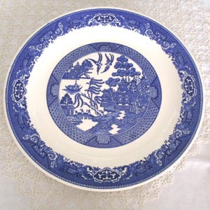May include: A white ceramic plate with a blue Willow pattern design. The plate features a central scene with a bridge, pagoda, and birds, surrounded by a detailed border. The plate is a classic piece of tableware.