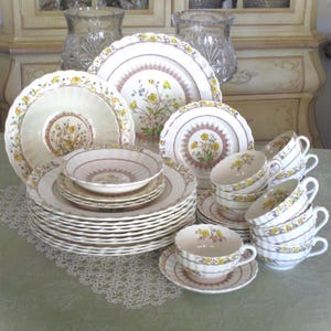 May include: A complete vintage floral china set featuring plates, bowls, and teacups. The white porcelain has a yellow floral design with a scalloped edge and a delicate pink border. Includes teacups, saucers, and serving dishes.