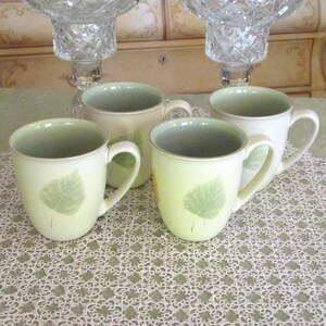 May include: Four cream-colored ceramic mugs with a green leaf design. The mugs have a light green interior and a curved handle. The mugs are on a white lace tablecloth. Two crystal candle holders are in the background.