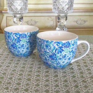 May include: Two white ceramic mugs with a blue floral pattern. The mugs have a rounded shape and a white handle. The floral design includes shades of blue, green, and purple. The mugs are on a white lace tablecloth.