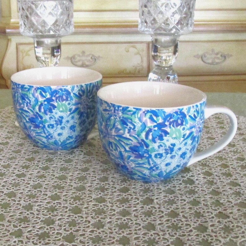 May include: Two white ceramic mugs with a blue floral pattern. The mugs have a rounded shape and a white handle. The floral design includes shades of blue, green, and purple. The mugs are on a white lace tablecloth.