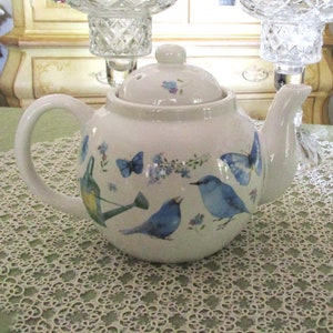 May include: White ceramic teapot with a bluebird and butterfly design. The teapot has a lid and a spout. The teapot is sitting on a white lace tablecloth.