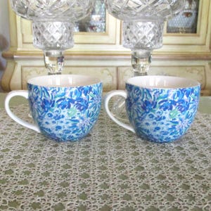 May include: Two ceramic mugs with white handles and a blue floral pattern. The mugs have a white interior and are sitting on a green lace tablecloth. Crystal candle holders are in the background.