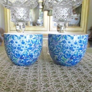 May include: Two matching decorative candle holders. Each has a clear, faceted crystal top and a blue and white floral patterned base. The bases are a rounded bowl shape. The background includes a lace tablecloth and a window.