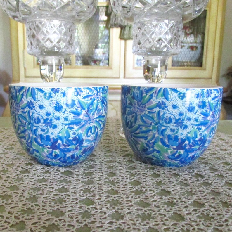 May include: Two matching decorative candle holders. Each has a clear, faceted crystal top and a blue and white floral patterned base. The bases are a rounded bowl shape. The background includes a lace tablecloth and a window.