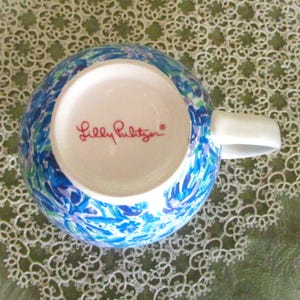 May include: A ceramic mug with a white handle and a vibrant blue, green, and purple floral pattern. The bottom of the mug features the text "Lilly Pulitzer" in red script.