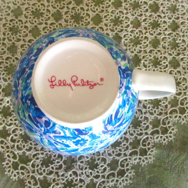 May include: A ceramic mug with a white handle and a vibrant blue, green, and purple floral pattern. The bottom of the mug features the text "Lilly Pulitzer" in red script.