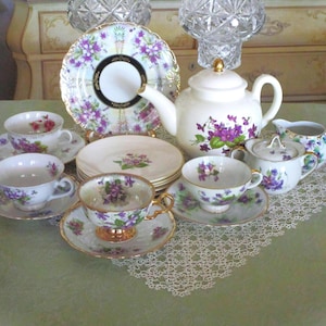 May include: A porcelain tea set featuring a white teapot, a creamer, a sugar bowl with lid, four teacups with matching saucers, and a decorative plate. Each piece is decorated with purple violet floral patterns and gold trim accents.
