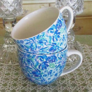 May include: Two stacked ceramic mugs with white handles and interiors. The exterior of each mug is decorated with a blue floral pattern on a white background. The mugs are sitting on a white lace tablecloth.
