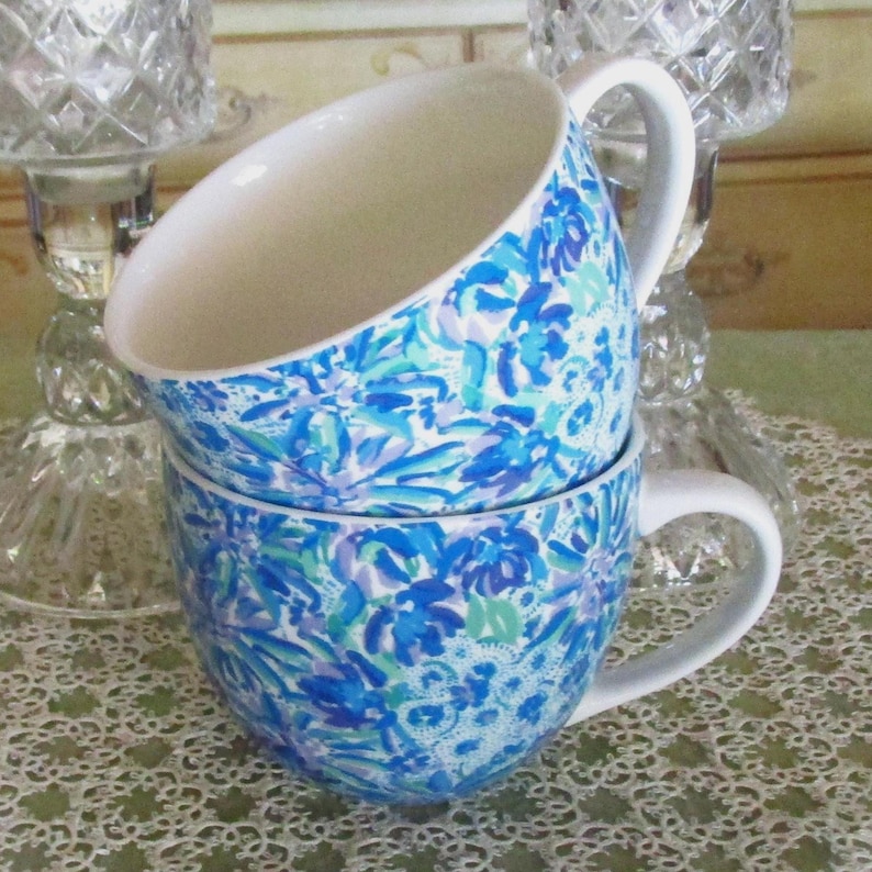 May include: Two stacked ceramic mugs with white handles and interiors. The exterior of each mug is decorated with a blue floral pattern on a white background. The mugs are sitting on a white lace tablecloth.