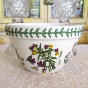 May include: A white ceramic planter with a green leaf border and floral and butterfly illustrations. The planter features a design of purple, yellow, and blue flowers and butterflies. The planter is sitting on a lace tablecloth.