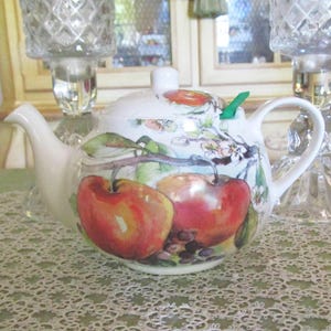 May include: White ceramic teapot with a lid, handle, and spout. The teapot is decorated with a watercolor painting of red apples, green leaves, and white blossoms. A green tea infuser is inserted into the spout.