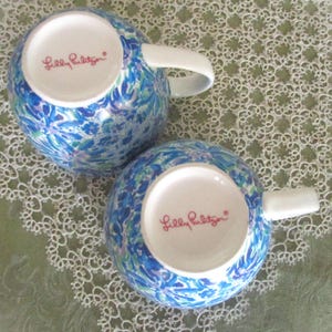 May include: Two round ceramic mugs with white handles and a blue floral pattern. The bottom of each mug has the words "Lilly Pulitzer" in pink script. The mugs are on a white lace tablecloth.