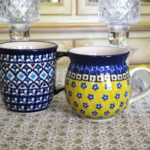 May include: Two ceramic mugs with intricate patterns. One mug is primarily blue with a geometric design, while the other is yellow with blue floral accents. Both mugs have a dark blue rim and handle.