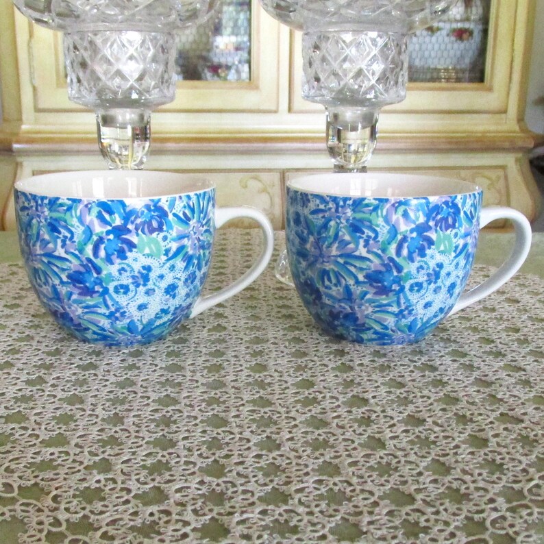 May include: Two ceramic mugs with white handles and a blue floral pattern. The mugs are placed on a white lace tablecloth. Above the mugs are two crystal candle holders.