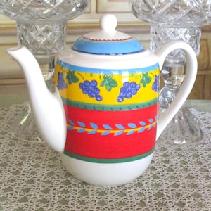 May include: A white ceramic teapot with a colorful design. The teapot features a blue lid, a yellow band with grape illustrations, and a red band with blue leaf accents. It has a curved spout and a handle.