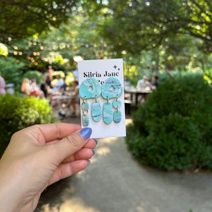 May include: A pair of teal blue and white marbled dangle earrings with gold accents. The earrings are displayed on a white card with the text "Silvia Jane" printed on it.