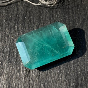 May include: A vibrant green gemstone with a faceted, rectangular shape. The gemstone is set against a dark gray background.