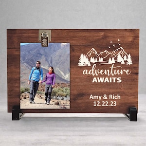 May include: A wooden photo frame with a picture of a couple hiking. The frame has the words "adventure awaits" and the names "Amy & Rich" with the date "12.22.23" printed on it. The frame is supported by two black metal stands.
