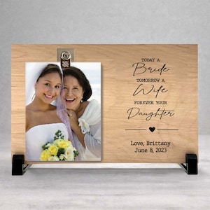 May include: A rectangular wooden photo display stand featuring a metal clip holding a photograph of a bride and an older woman. The wood is engraved with the text: TODAY A Bride TOMORROW A Wife FOREVER YOUR Daughter Love, Brittany June 8, 2023.