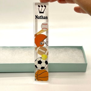 May include: A clear acrylic mezuzah with a sports theme. The mezuzah has a baseball, football, soccer ball, tennis ball, and basketball design. The name "Nathan" is printed on the mezuzah.