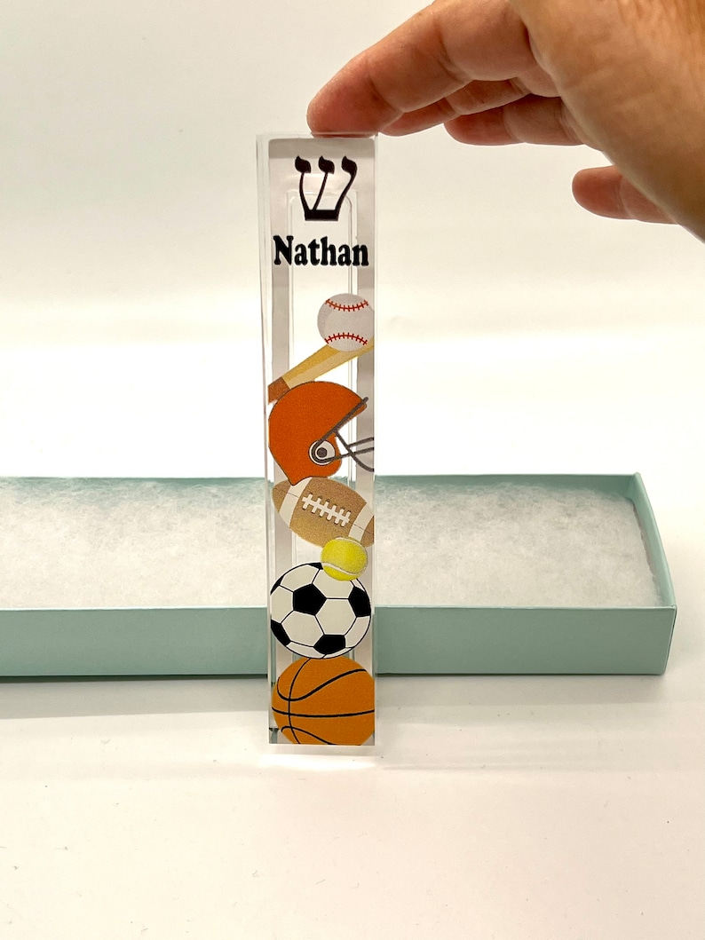 May include: A clear acrylic mezuzah with a sports theme. The mezuzah has a baseball, football, soccer ball, tennis ball, and basketball design. The name "Nathan" is printed on the mezuzah.