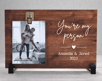 May include: A rectangular dark brown wood photo frame featuring a metal clip at the top. The frame displays a photograph of two people and the white printed text "You're my person" above a heart, "Amanda & Jared", and "2023".