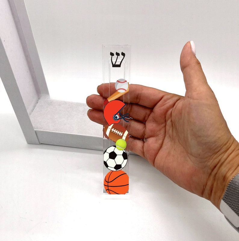 May include: A clear acrylic sports themed mezuzah with a baseball, football, basketball, tennis ball, and a baseball bat. The mezuzah has the Hebrew letter Shin on the top.