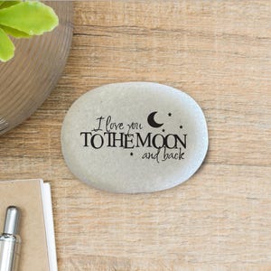 May include: A smooth, light gray oval stone featuring black printed text that reads I love you TO THE MOON and back, accompanied by a small crescent moon icon and three tiny star icons positioned above and below the central lettering.