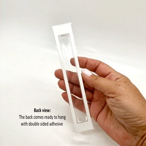 May include: White plastic frame with a clear plastic insert. The back of the frame has double-sided adhesive for hanging. Back view: The back comes ready to hang with double sided adhesive.