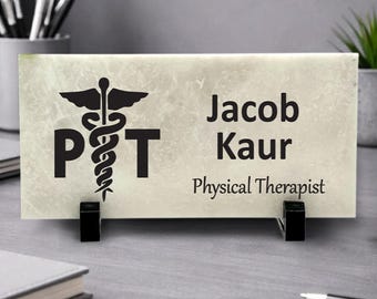 May include: A rectangular desk nameplate made of light-colored marble with black text and graphics. The left side features a black caduceus symbol between the letters P and T. The right side displays the name Jacob Kaur and Physical Therapist.