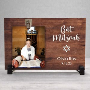 May include: A wooden photo frame with a picture of a Bat Mitzvah ceremony. The frame has the words "Bat Mitzvah", a Star of David, the name "Olivia Ray", and the date "9.18.23" in white script. The frame is supported by black stands.