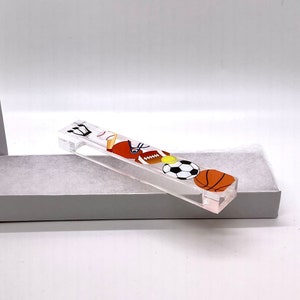 May include: A clear acrylic sports themed mezuzah with a baseball, football, soccer ball, and basketball design.