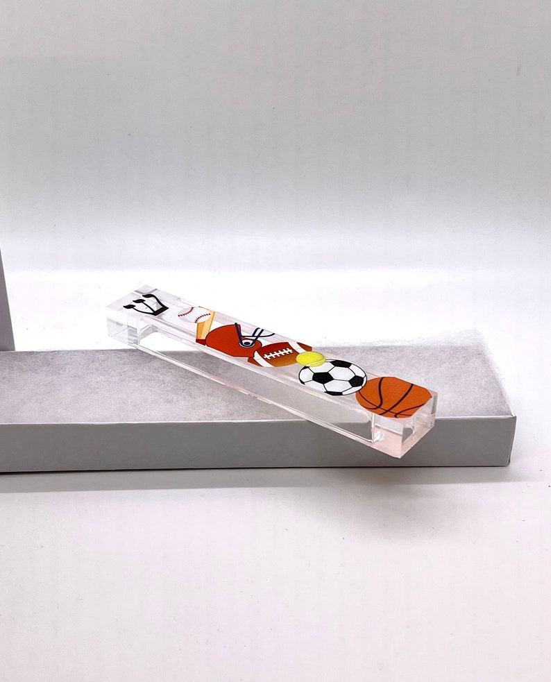 May include: A clear acrylic sports themed mezuzah with a baseball, football, soccer ball, and basketball design.