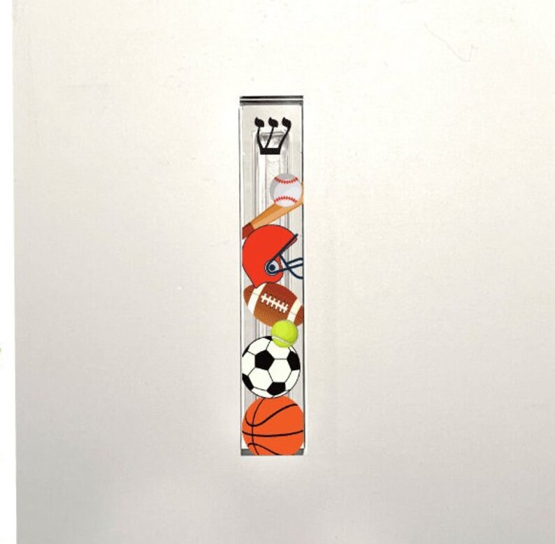 May include: A clear acrylic mezuzah with a sports theme. The mezuzah features a baseball, football, basketball, soccer ball, and a baseball bat. The mezuzah is decorated with a Hebrew letter Shin.