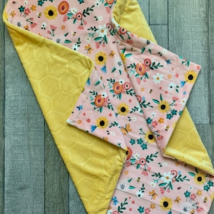 May include: A baby blanket with a floral pattern and a yellow honeycomb design. The blanket features a pink background with sunflowers, bees, and other flowers. The reverse side is a soft yellow fabric.