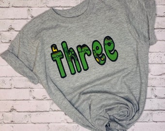 Puede incluir: Una camiseta gris con la palabra "Three" en letras grandes y verdes con un diseño de tractor. La camiseta está sobre un fondo de pared de ladrillos blancos.