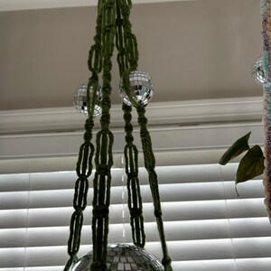 May include: A green macrame plant hanger with three silver disco ball ornaments. The hanger is hanging from a ceiling and the disco balls are at different heights.