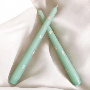 May include: Two seafoam green taper candles with seashell and starfish designs. The candles are crossed, creating an X shape. The candles are decorated with small, light pink seashell and starfish illustrations.